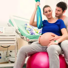 Pareja en sala de partos. Ella sentada en balón, buscando la mejor postura para parir, él la abraza.
