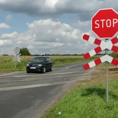 Czarny samochód zbliża się do przejazdu kolejowego z czerwonym znakiem STOP i białoczerwonym krzyżem.
