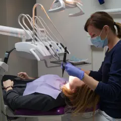 Dentystka w masce i rękawiczkach pracuje przy pacjencie. W gabinecie widoczny sprzęt do stomatologii.