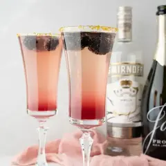 Dwa eleganckie drinki z prosecco, ozdobione jeżynami i złotym cukrem, obok butelek prosecco i wódki.