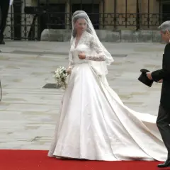 Kate Middleton w drugiej sukni ślubnej, z welonem i bukietem, idzie po czerwonym dywanie. Obok niej mężczyzna w żakiecie trzyma cylinder.