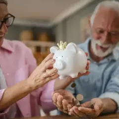 Uśmiechnięta para seniorów wsypuje monety do skarbonki-świnki. Czekają na moment, kiedy wyrównają emerytury czerwcowe.