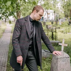 Mężczyzna w żałobnym stroju pochyla się nad kamiennym nagrobkiem z krzyżem na cmentarzu.