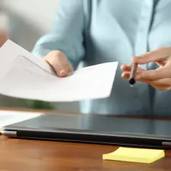 Dłoń podaje dokument, sugerując omawianie różnych form zatrudnienia. Na biurku leży laptop i żółta karteczka.