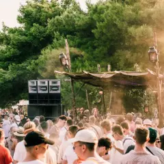 Folla in delirio a un festival tecno all'aperto, con enormi casse acustiche e un palco improvvisato tra gli alberi.