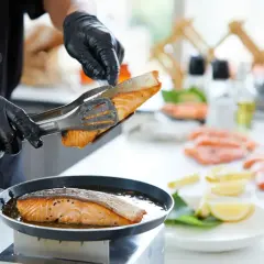 Szef kuchni w czarnych rękawiczkach kładzie łososia na patelni. Smażenie ryby na złoto to właśnie sauté.