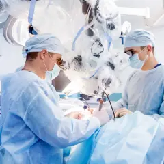 Chirurdzy w maseczkach i fartuchach operacyjnych pracują przy użyciu mikroskopu.