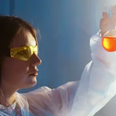 Naukowczyni w okularach ochronnych analizuje płyn w kolbie. To jej praca w dziedzinie biotechnologii.