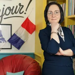 Kobieta w okularach stoi przed ścianą z napisem "Bonjour!", francuską flagą i wieżą Eiffla.