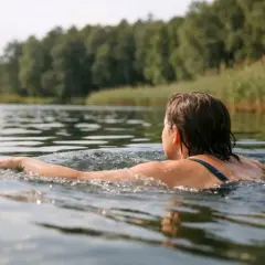Kobieta pływa w jeziorze, ciesząc się letnim dniem. Woda jest spokojna, a na horyzoncie widać las.