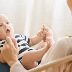 Ein Baby mit offenem Mund liegt auf dem Schoß seiner Mutter. Es hält ihre Hände. Bald sagt das Baby "Mama".
