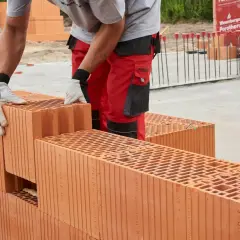Budowa domu z pustaków ceramicznych Porotherm. Pracownik w czerwonych spodniach układa cegły, pokazując, z czego budować dom.