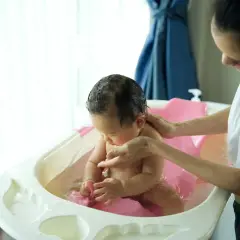 Mama delikatnie kąpie dziecko w wanience. Woda jest ciepła, a maluch bawi się zabawką. Może dodano krochmal do kąpieli dla miękkości skóry?