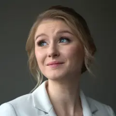 Portret młodej kobiety z blond włosami i niebieskimi oczami, w białej marynarce. Jej wiek to około 30 lat.