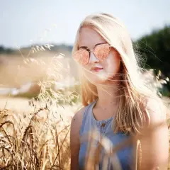 Dziewczyna z piaskowym blond włosami w okularach przeciwsłonecznych stoi w złotym polu pszenicy.
