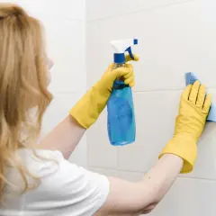 Kobieta w żółtych rękawiczkach używa środka do czyszczenia, by usunąć pleśń w łazience.