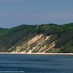 Zielone wzgórza porośnięte lasem schodzą ku morzu, odsłaniając piaszczystą, białą górę. Na szczycie majaczy ceglana wieża.
