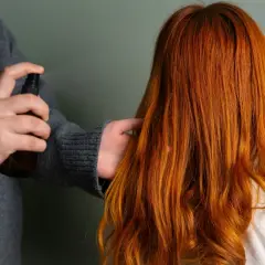 Persona rocía un producto en el cabello fino y quebradizo de color naranja rojizo para darle brillo y protección.