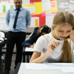 Dziewczynka z ołówkiem w ustach pisze test, nauczyciel stoi z tyłu.