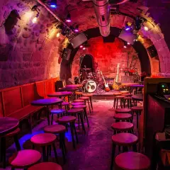 Scène de concert dans le caveau des oubliettes, éclairée par des lumières violettes et rouges. Tables et tabourets disposés devant la scène où se trouve une batterie et des guitares.