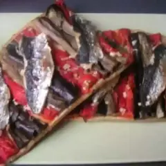 Deux tartes rectangulaires de coca catalane garnies de poivrons rouges, d'aubergines et de sardines, sur une assiette verte.