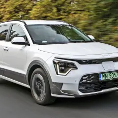 Biały Kia e-Niro jedzie po drodze, otoczony zielenią drzew.