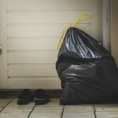 A full black trash bag and a pair of slippers sit by a closed door, symbolizing the need for lean budgeting to discard unnecessary expenses.