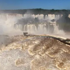 Mächtige Iguazú-Wasserfälle stürzen in schlammbraunes Wasser. Ein Steg führt durch den Sprühnebel, ein Regenbogen funkelt.