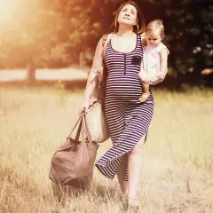 Schwangere Frau mit Baby im Arm und Taschen, die sich durch hohes Gras kämpft. Schwer heben schwangerschaft.