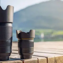 Two camera lenses, a tall telephoto and a shorter normal lens, sit on a wooden deck with a blurred mountain and lake background.