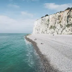 Plage de galets de la Baie de Somme, avec des falaises blanches imposantes et une mer d'un bleu-vert apaisant. Quelques personnes profitent du soleil.