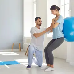Fizjoterapeuta pomaga kobiecie w ćwiczeniu aktywującym mięśnie głębokie przy użyciu piłki gimnastycznej.
