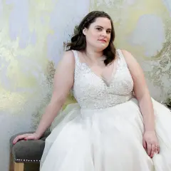 Kobieta w białej, zdobionej sukni ślubnej plus size siedzi na tle złoto-białej ściany.