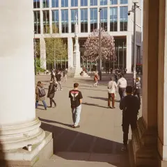 A 35mm street photography scene captures people walking in a plaza with a modern building and blooming trees.