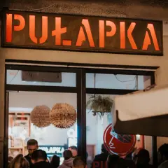 Neonowy napis "PUŁAPKA" nad wejściem do lokalu. W środku widać ludzi i dekoracje.