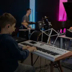 Chłopiec na lekcjach śpiewu ćwiczy grę na keyboardzie, podczas gdy w tle słychać perkusję i mikser dźwięku.