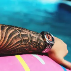 Ręka z tatuażem w stylu tribal, zegarek na nadgarstku, leżąca na dmuchanym kole w basenie. Idealny tatuaż w lato.