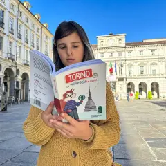 Bambina legge una guida "Torino con Viaggiapiccoli" in una piazza torinese. La libreria viaggi è perfetta per scoprire la città.