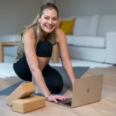 Glad kvinna förbereder sig för kundaliniyoga positioner hemma, med en laptop och yogablock redo.