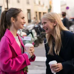 Camille och Emily i Paris, skrattandes med kaffe och blommor på en solig gata.