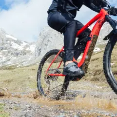 Kolarz na czerwonym rowerze trekkingowym pokonuje błotnistą kałużę na górskim szlaku.
