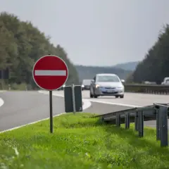 Czerwony znak z białym pasem informuje o zakazie wjazdu. Droga zakręca, a w oddali widać jadące samochody.
