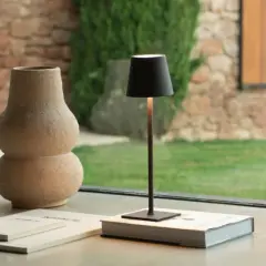 Elegantes ideas de lámparas para tu hogar: una lámpara negra minimalista sobre un libro, junto a un jarrón texturizado y un jardín exterior.