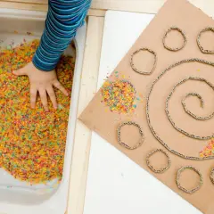 Main colorée dans un bac, une main d'enfant explore. À côté, un carton en spirale avec des ronds découpés, parsemé de riz coloré. Une activité sensorielle adulte pour stimuler la créativité.