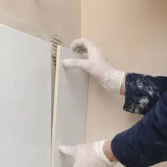 Mani guantate applicano una piastrella bianca su un muro, mostrando come attaccare piastrelle al muro con precisione.
