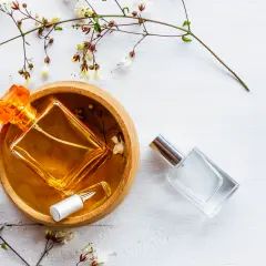 Dwie buteleczki perfum, jedna z bursztynowym płynem w drewnianej miseczce, druga szklana. Delikatne gałązki z kwiatami. Woda toaletowa ile alkoholu?