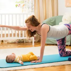 Mama wykonuje ćwiczenia po porodzie na macie, a jej maluch leży obok.