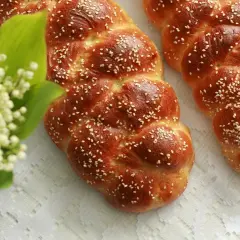 Dwie złociste chały posypane sezamem, obok bukiecik konwalii na białym obrusie.