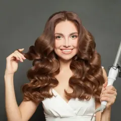 Uśmiechnięta kobieta z falowanymi brązowymi włosami, prezentująca lokówkę. Efektowne styling włosów.