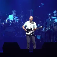 Mark Knopfler na scenie z zespołem podczas jednego z jego niezapomnianych koncertów. Muzycy grają na gitarach, saksofonach i perkusji.